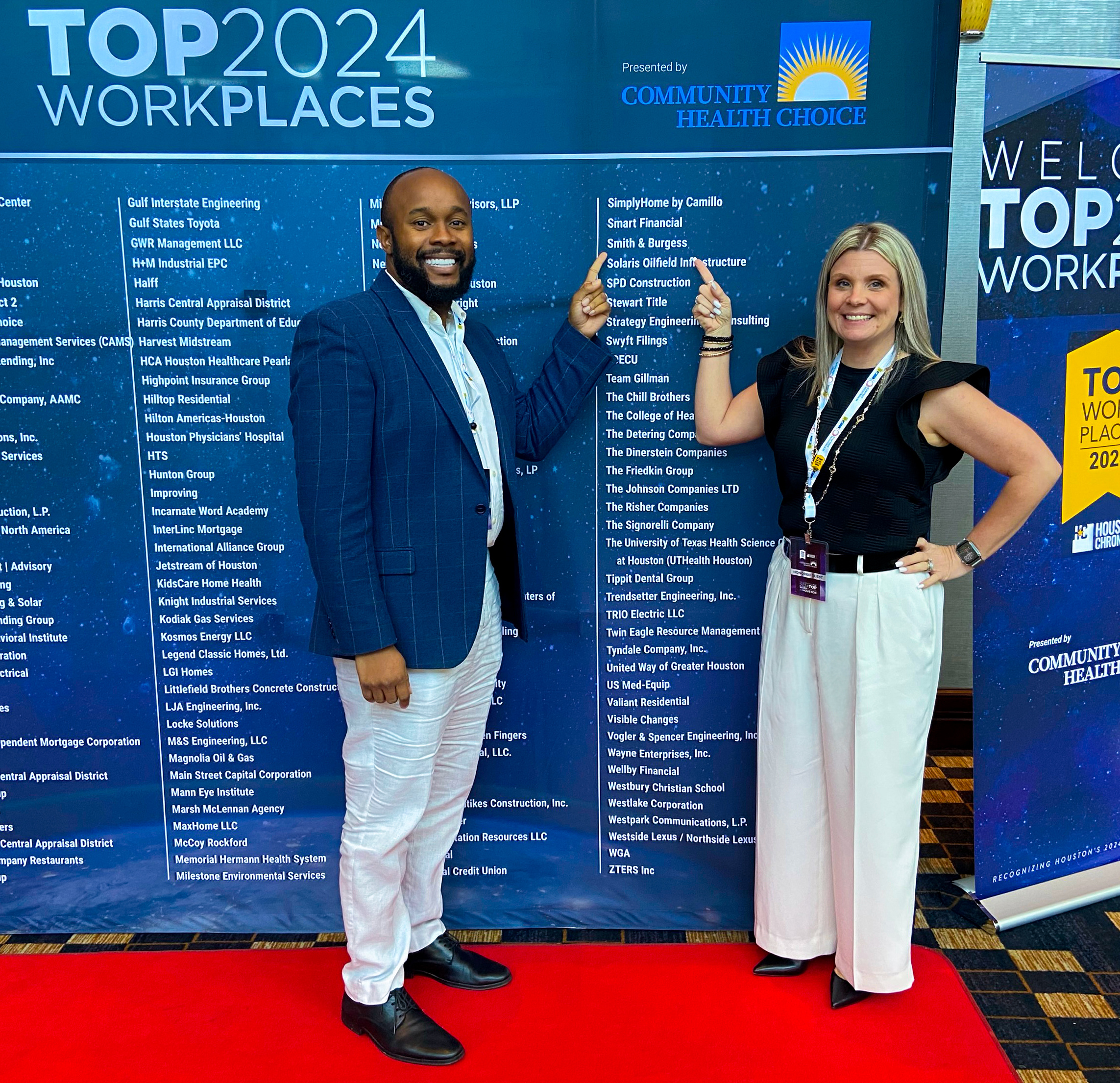 Two Smith & Burgess employees standing on a red carpet, smiling and pointing to the company’s name listed on the Houston Chronicle’s Top Workplaces 2024 display banner.